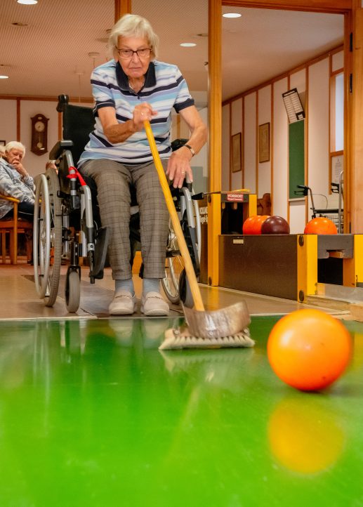 Dame spielt kegeln mit einer Bowlingkugel und einem Besen im Rollstuhl
