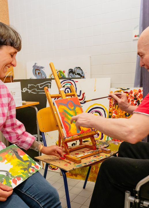 Ein Herr ist mit einer Betreuerin im Atelier und bemalt eine Leinwand